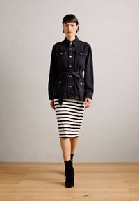 Veste en denim noire avec des boutons et une ceinture à la taille, assortie à une jupe crayon noire et blanche à rayures serrées, et des bottines.