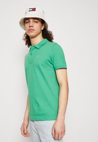 Polo shirt verde in cotone con colletto, maniche corte e piccolo logo ricamato sul petto, abbinato a un cappello a secchiello bianco.