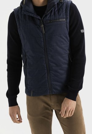 Mężczyzna ubrany w granatową pikowaną kamizelkę na czarnym swetrze z długim rękawem i brązowe spodnie, stoi na tle jednolitego jasnego tła.