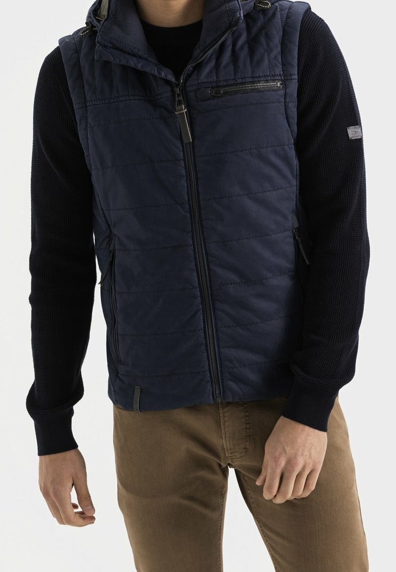 Homme portant un gilet matelassé bleu marine sur un pull à manches longues noir avec un pantalon marron, debout devant un fond clair uni.