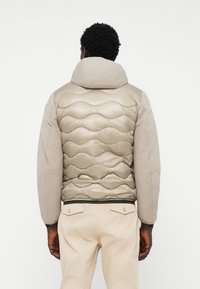 Gevoerde jas in gedempt beige met een gewatteerd golfpatroon op de rug, voorzien van een capuchon en soepele mouwen, afgewerkt met een geribbelde tailleband.