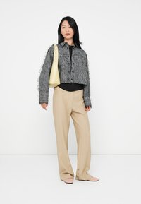 Jonge vrouw in een beige broek, met een gestructureerde grijze button-up jas, die een lichtgele schoudertas draagt en lichte roze schoenen aan heeft.