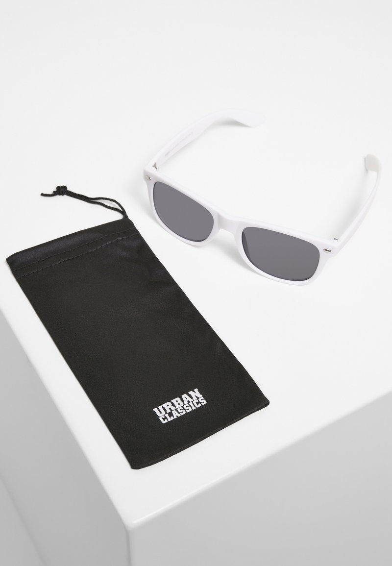 Ochelari de soare din plastic alb cu lentile gri, lângă un pachet din material textil negru, având un logo alb „URBAN CLASSICS” cu litere îngroșate.