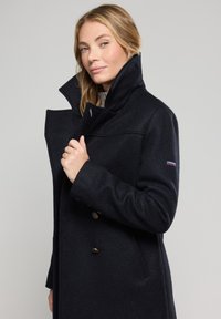 Abrigo de lana azul marino con un alto cuello y diseño de doble botonadura. Presenta grandes botones circulares y un pequeño logotipo en la manga.