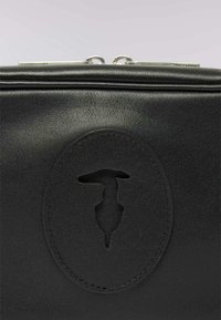 Svart läderväska med en slät yta, med en cirkulär präglad logotyp i mitten och silverfärgad hårdvara vid toppdragkedjan.