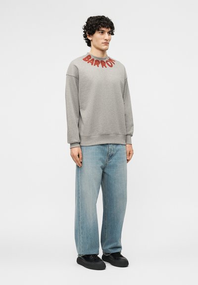 Άνδρας που φοράει ανοιχτό γκρι φούτερ με τη λέξη "BARROW" σε κόκκινα γράμματα στο γιακά, φαρδιά μπλε τζιν και μαύρα αθλητικά παπούτσια, στέκεται μπροστά σε λευκό φόντο.