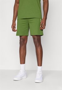 Calções de algodão verdes com uma cintura elástica, bolsos laterais e um ajuste slim. Combinados com ténis brancos e meias de cano curto.