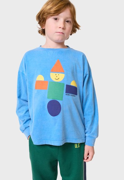 Vaaleansininen pitkähihaista T-paitaa, jossa on värikäs geometrinen kuvio ja hymyilevä hahmo. Yhdistetty vihreisiin housuihin, joissa on keltainen "B.C."