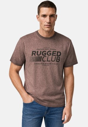 Homme portant un t-shirt brun "Rugged Club" avec texte, jean bleu, debout avec une main dans la poche sur un fond uni.