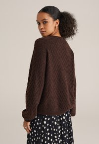 Rjav pleteni pulover z vzorcem ribje kosti, kombiniran s črno krilo z belimi pikami.