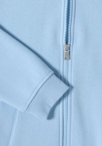 Sweat-shirt zippé bleu clair avec fermeture éclair métallique visible et poignet côtelé sur la manche longue.