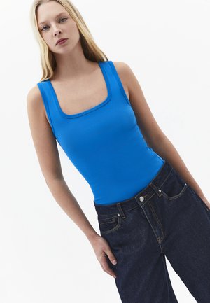 Jeune femme aux longs cheveux blonds portant un haut sans manches bleu vif et un jean taille haute foncé sur un fond blanc uni.