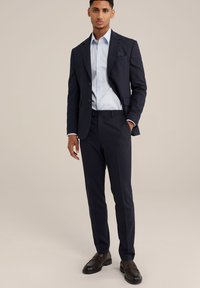 Marineblauw op maat gemaakt pak met een gestructureerde afwerking, aangevuld met een lichtblauwe blouse. Bruine loafers maken de formele uitstraling compleet.