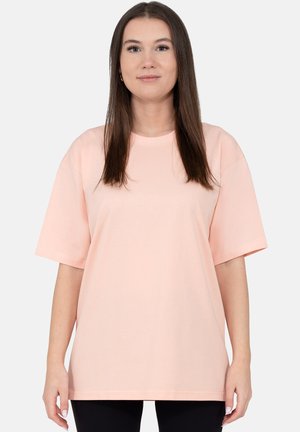 Młoda kobieta z długimi brązowymi włosami, ubrana w luźną jasnoróżową koszulę z krótkim rękawem na jednolitym tle.