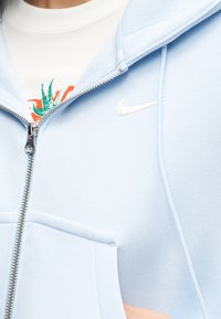 Κοντινό πλάνο ενός ατόμου που φοράει ένα ανοιχτό μπλε φούτερ Nike με φερμουάρ πάνω από ένα λευκό παντελόνι με πράσινο και πορτοκαλί γραφικό σχέδιο.