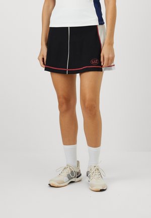 Black sporty skirt with red and white accents, featuring a logo on the front and contrasting stitching. Worn with white socks and beige shoes.
