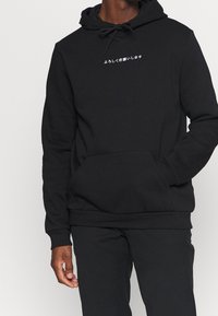 Felpa nera con cappuccio in misto cotone. Presenta una tasca a marsupio, cappuccio con coulisse e testo bianco in giapponese sul petto.