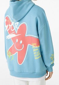 Sudadera azul clara con una gran estrella sonriente rosa, texto amarillento escrito a mano y diseños de estrellas en la manga, vista desde la parte posterior de una persona.