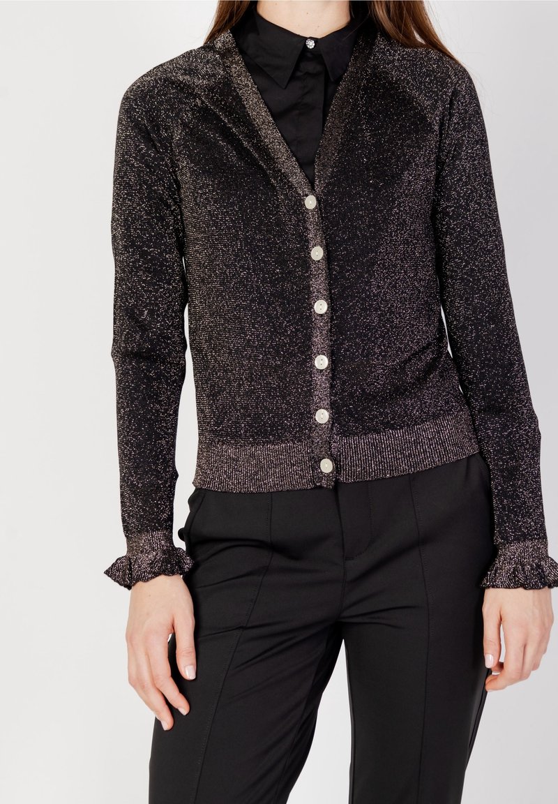 Cardigan noir avec un subtil éclat, doté d'un col en V, de six boutons et de poignets volants. Porté sur une chemise noire et un pantalon ajusté.