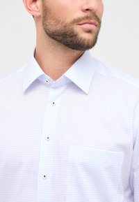 Uomo con barba che indossa una camicia a quadri leggera blu chiaro con taschino, colletto e bottoni bianchi, mentre guarda di lato.