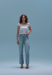 Off-shoulder hvit topp kombinert med lyseblå høytlivs jeans og svarte sandaler med høye hæler, foran en blå bakgrunn.
