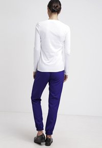 Weißes Langarmoberteil und lila Sweatpants mit elastischen Bündchen; Model trägt schwarze Slipper, von hinten betrachtet.