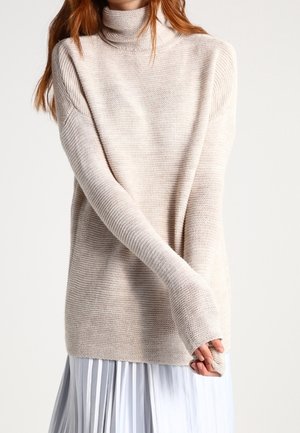 Frau trägt einen beigen, gerippten Rollkragenpullover mit langen Ärmeln über einem weißen, plissierten Rock, die Arme verschränkt und die Hände teilweise bedeckt.