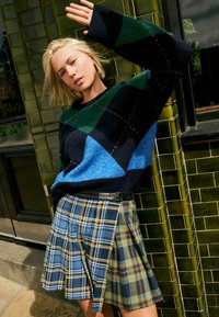 Pull à motifs marine, vert et bleu avec des losanges ; jupe à carreaux dans des tons jaunes, bleus et verts ; coupe ajustée et détail de ceinture.