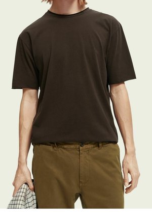 Person trägt ein einfarbiges dunkelbraunes kurzärmliges T-Shirt und braune Hose, hält ein kariertes Tuch in der linken Hand.
