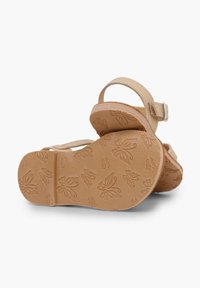 Beige sandaalit matalalla korolla, joissa on säädettävä nilkkaremmi. Ulkopohjassa on kohokuvioitu perhoskuvio koristeluna.