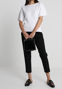 Top blanco de manga corta con escote redondeado, combinado con pantalones negros ajustados. La modelo sostiene un pequeño bolso negro y lleva zapatos negros sin cordones con adornos.