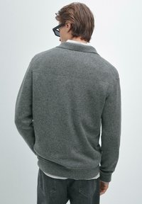 Uomo che indossa un maglione grigio scuro, una camicia bianca e jeans neri, di spalle contro uno sfondo grigio chiaro uniforme.