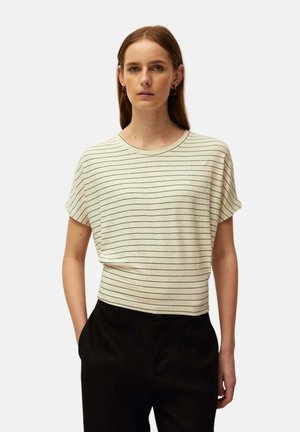Mujer lleva una camiseta beige de manga corta con finas rayas horizontales negras y pantalones negros de cintura alta, de pie con las manos en los bolsillos.