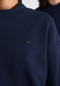 Nahaufnahme eines navyblauen Sweatshirts mit einem gestickten dunklen Krokodil-Logo auf der Brust.