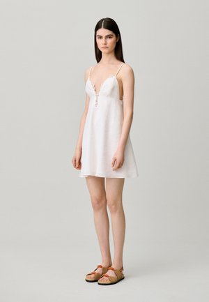 Joven mujer con el pelo largo y oscuro, vestida con un vestido blanco sin mangas con detalles de encaje y sandalias beige con tiras naranjas, de pie sobre un fondo gris.