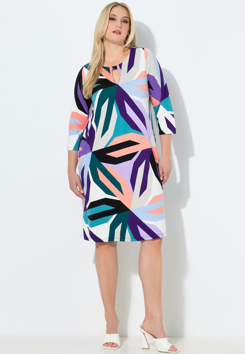 Nainen polvipituinen mekko, jossa abstrakti monivärinen geometrinen kuvio, neljänneshihat ja valkoiset korolliset sandaalit, seisomassa valkoista taustaa vasten.