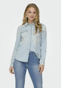 Camicia di jeans azzurro chiaro con maniche lunghe, due tasche sul petto, bottoni a pressione e orlo curvo, abbinata a jeans blu. Tessuto liscio.