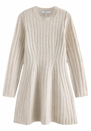 Kobieta w beżowej, prążkowanej dzianinowej sukience z długim rękawem, białych skarpetkach i czarnych mokasynach, stojąca na tle jasnobeżowego tła.