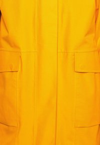 Impermeabile giallo brillante con un design elegante, dotato di due tasche frontali e cuciture a contrasto, realizzato in materiale resistente all'acqua.