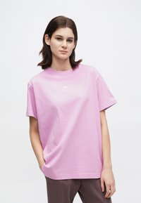 Noor, kellel on tume juuksed, kannab lohakat heleda lillat Nike T-särki ja tumedaid pükse, seistes neutraalse heleda tausta ees.