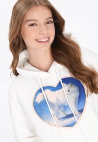 Fehér kapucnis pulóver, amelyen szív alakú grafika látható egy fehér állattal kék háttér előtt. Texturált anyag, húzózsinóros kapucni és hétköznapi kialakítás.