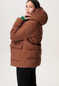 Bruine pufferjack met capuchon, uitgerust met een gestikte uitstraling, zijzakken en een zachte textuur. Model draagt een groene trui eronder.