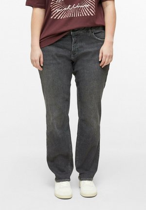 Person trägt dunkelgraue Jeans, weiße Turnschuhe und ein weinrotes T-Shirt mit grafischem Design und steht vor einem schlichten Hintergrund.