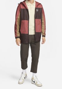 Bunda s bloky barev v barvách burgundské červené, olivové a hnědé, s uzavíráním na zip, kapucí a dvěma předními kapsami. Nošena přes béžové polo tričko.