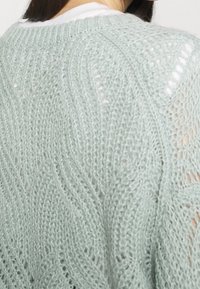 Pull en tricot vert clair avec un motif de vagues et une texture ajourée. Fil doux et coupe décontractée, avec un col côtelé.