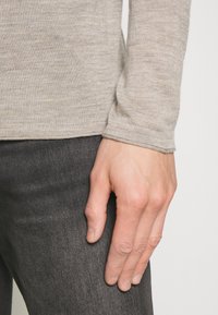 Beiges Langarmshirt aus strukturiertem Strickstoff, kombiniert mit dunklen Denim-Jeans. Nahaufnahme einer Hand, die am Oberschenkel ruht.