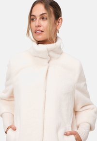 Manteau en peluche couleur crème avec un col montant, des manches larges et des poignets en tricot côtelé. Doté d'une fermeture à l'avant et de poches latérales pour plus de praticité.