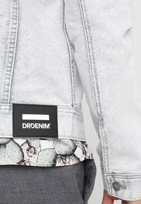 Veste en denim délavé avec étiquette noire "DRDENIM" au dos, portée sur une chemise à motif cactus et un pantalon gris.
