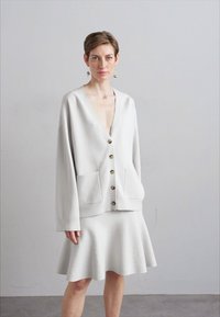 Γυναίκα με κοντά μαλλιά φοράει υπερμεγέθη ανοιχτό γκρι πουλόβερ με κουμπιά και μεγάλες τσέπες, συνδυασμένο με φούστα που φαρδαίνει προς τα κάτω, σε στάση απέναντι από απλό τοίχο.