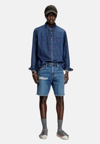 Camicia di jeans con maniche lunghe e colletto, abbinata a pantaloncini di jeans strappati. Calzini grigi e scarpe grigie con dettagli rossi completano il look.